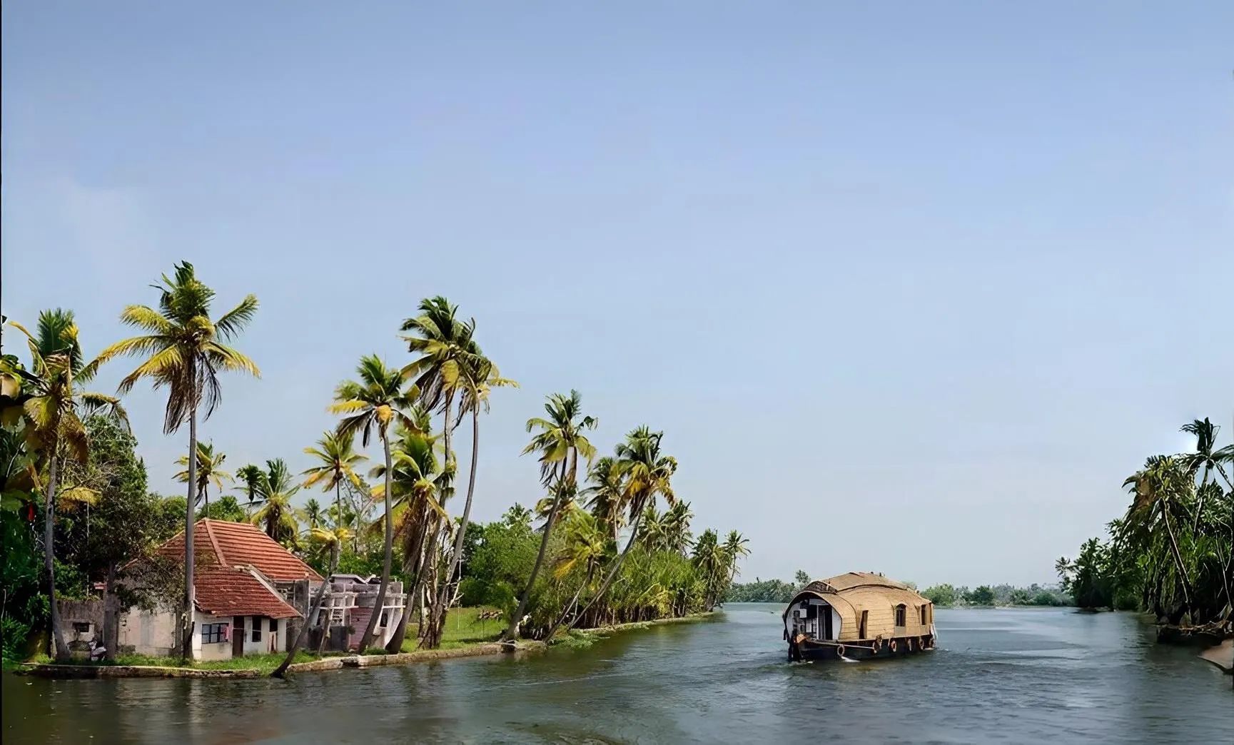 A traditional houseboat cruising through the serene backwaters of Kakkathuruth in Alappuzha, Kerala, surrounded by lush coconut palm trees and village houses under a clear blue sky.